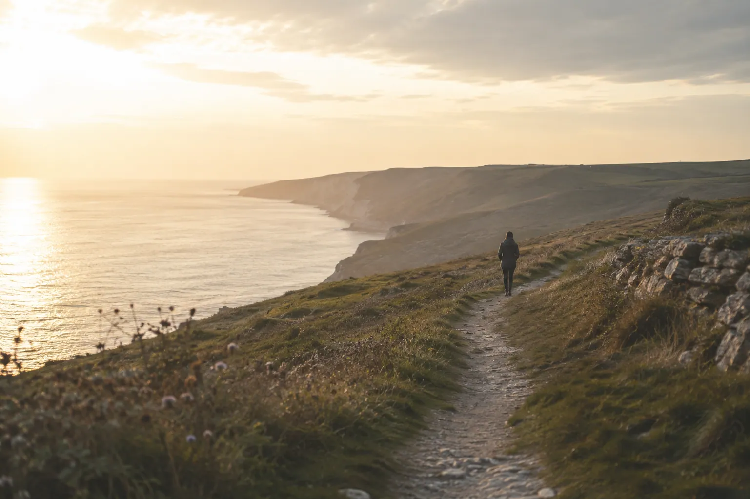 A person walking a footpath along a grassy clifftop above the sea at sunset